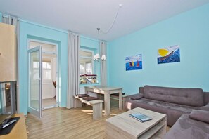 A modern living room with a sofa, coffee table, and a dining area with a table and chairs. There is a window with curtains, a painting on the wall, and a TV on the left. - Quiet location, beach-style vacation apartment with a veranda - Villa Möwe FeWo 06 - Villa (Göhren)