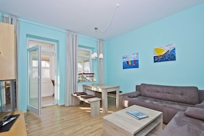 A modern living room with a sofa, coffee table, and a dining area with a table and chairs. There is a window with curtains, a painting on the wall, and a TV on the left. - Quiet location, vacation apartment in seaside style with veranda - Villa Möwe FeWo 06 - Villa (Göhren)