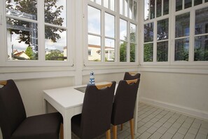 A room with a white table and four chairs, large windows with white frames, and a view of houses and trees outside. - Quiet location, porch, free use of the AHOI adventure pool - Villa Möwe FeWo 01 - (Göhren)