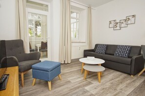 A modern living room with a grey sofa, a blue ottoman, and a white coffee table. - Quiet location, porch, free use of the AHOI adventure pool - Villa Möwe FeWo 01 - (Göhren)