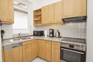 A compact kitchen with wooden cabinets, a built-in microwave, a coffee maker, and a stove. - Quiet location, porch, free use of the AHOI adventure pool - Villa Möwe FeWo 01 - (Göhren)