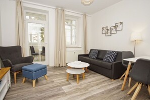 A modern living room with a grey sofa, blue ottoman, white coffee table, and a small white table with a lamp. - Quiet location, porch, free use of the AHOI adventure pool - Villa Möwe FeWo 01 - (Göhren)
