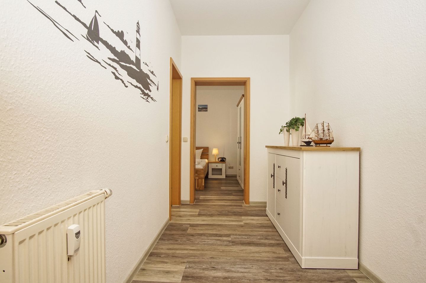 A narrow hallway with a wooden floor, a radiator on the left, a white cabinet on the right, and a door at the end leading to a room with a bed and a nightstand.