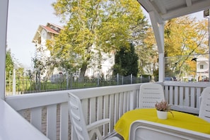 A covered outdoor seating area with white chairs and a table, a potted plant, and a view of a tree-lined street. - Quiet location with a terrace; access to the pool and sauna - Villa Käthe FeWo 04 - (Göhren)