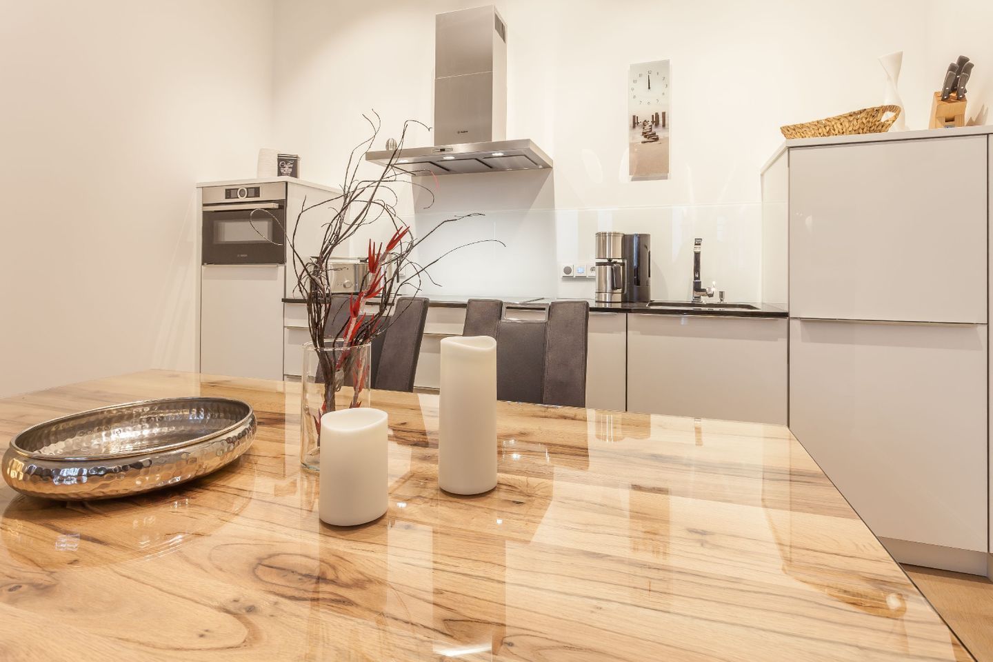 A modern kitchen with a wooden table, a stainless steel bowl, two white candles, and a vase with branches.