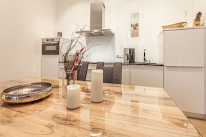 A modern kitchen with a wooden table, a stainless steel bowl, two white candles, and a vase with branches.