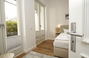 A bedroom with a bed, a bedside table, a nightstand, a window with blinds, and a door with a handle.