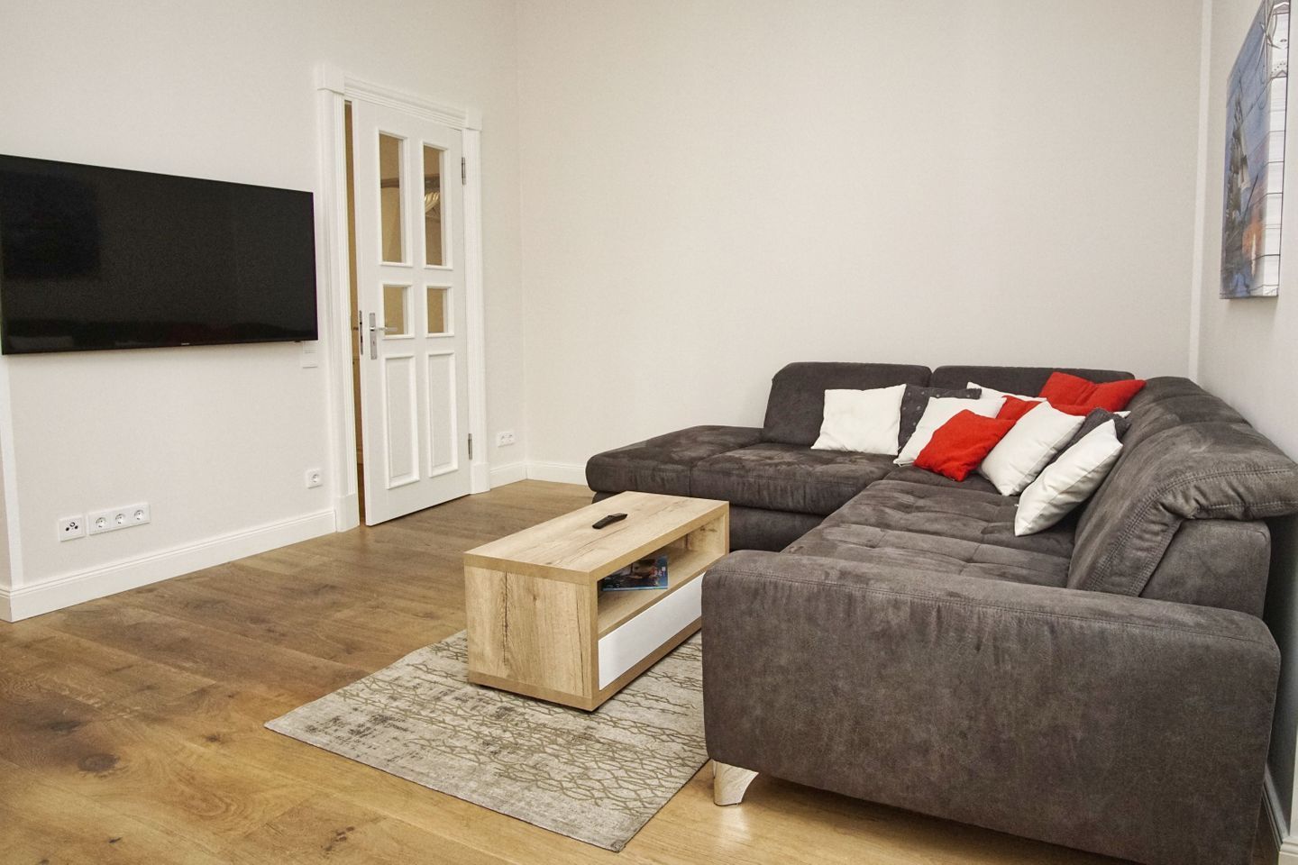 A living room with a grey sofa, a wooden coffee table, a flat-screen TV mounted on the wall, and a white door.