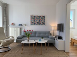 Ein modernes Wohnzimmer mit einem grauen Sofa, zwei weißen Beistelltischen, einem beigen Sessel, einem Fernseher und einer Heizung.