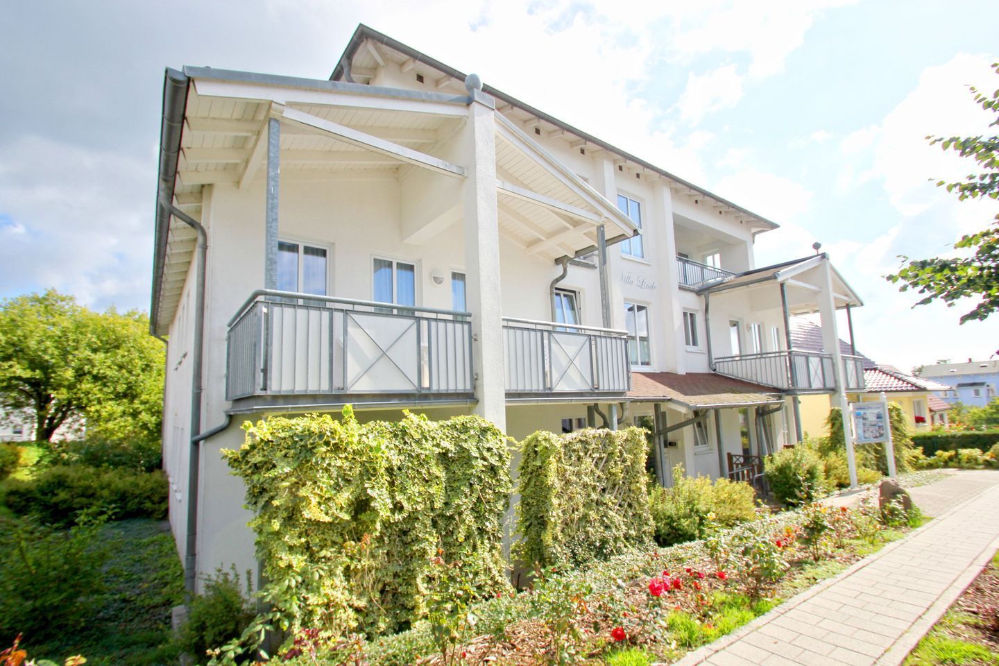 A multi-story building with balconies, a covered porch, and a garden with flowers and shrubs.