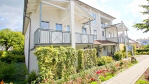Un immeuble à plusieurs étages doté de balcons, d’un porche couvert et d’un jardin agrémenté de fleurs et d’arbustes.