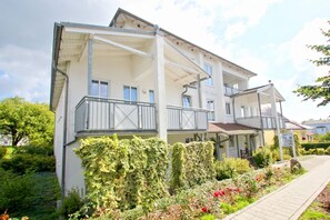 Un immeuble à plusieurs étages doté de balcons, d’un porche couvert et d’un jardin agrémenté de fleurs et d’arbustes.
