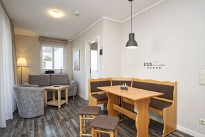 A modern, well-lit room with a sofa, a small table, and a wooden bench with cushions. There is a wall-mounted sign that reads "ESS Klasse" with three stars. - Die Ferienwohnung Nr. 27 in der Villa Buskam - Villa Buskam (Göhren)