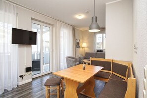 A modern living room with a wooden table, two chairs, a sofa, a TV mounted on the wall, and a ceiling light. - Die Ferienwohnung Nr. 27 in der Villa Buskam - Villa Buskam (Göhren)