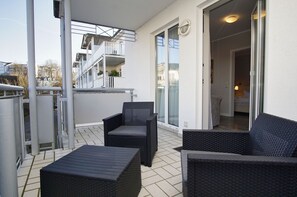 Ein Balkon mit Korbmöbeln, eine Glastür zu einem Innenraum und Blick auf ein Gebäude mit Balkonen.