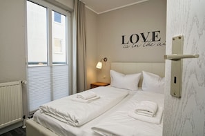Camera d'albergo con due letti, un muro con un cartello che recita 'LOVE is in the air', una finestra con persiane e un comodino con una lampada.
