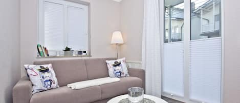 A living room with a beige sofa, two decorative pillows, a small side table with a glass vase, and a window with blinds.