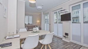 A modern dining area with a wooden table, white chairs, and a pendant light. A television is mounted on the wall, and there are windows with blinds.