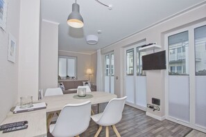 Ein moderner Essbereich mit einem Holztisch, weißen Stühlen und einer Pendelleuchte. Ein Fernseher ist an der Wand befestigt, und es gibt Fenster mit Jalousien.