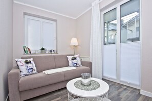Sala de estar com sofá bege, duas almofadas decorativas, mesa lateral pequena com um vaso de vidro e janela com persianas.