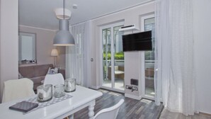 A modern living room with a white sofa, a dining table with a glass top, and a large TV mounted on a wall.