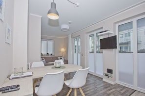 A modern dining area with a wooden table, white chairs, and a pendant light. A television is mounted on the wall, and there are windows with blinds. - Die Ferienwohnung Nr. 22 in der Villa Buskam - Villa Buskam (Göhren)