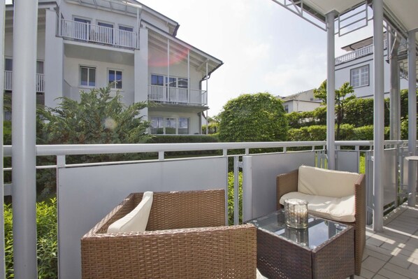 A balcony with wicker furniture and a glass table, overlooking a residential area with white buildings and greenery.