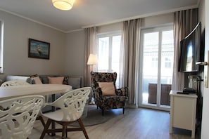 A modern living room with a sofa, a floral armchair, a dining table with chairs, a TV, and a large window with curtains. - Die Ferienwohnung Nr. 28 in der Villa Buskam - Villa Buskam (Göhren)