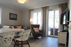 Ein modernes Wohnzimmer mit einer Couch, einem blumigen Sessel, einem Esstisch mit Stühlen, einem Fernseher und einem großen Fenster mit Vorhängen.