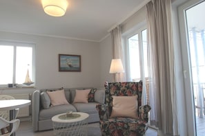 A living room with a grey sofa, a floral armchair, a round coffee table, and a painting of a ship on the wall. - Die Ferienwohnung Nr. 28 in der Villa Buskam - Villa Buskam (Göhren)