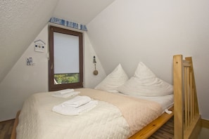 Een kleine, driehoekige kamer met een bed, kussens, een houten hoofdbord, een raam met jaloezieën en een klein wandklokje.