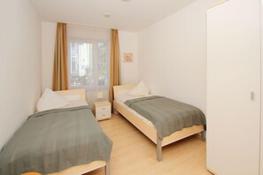 Huoneessa on kaksi erillistä sänkyä, pieni yöpöytä lampun kanssa, ikkuna, jossa on ohuet verhot, ja ovi.