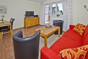 A living room with a red sofa, a wooden coffee table, a TV on a wooden cabinet, and a dining area with chairs and a table. - Central but quiet, balcony, free swimming pool and sauna - Villa Elisenhof apartment 2 (Göhren)
