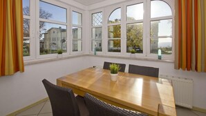 A dining area with a wooden table, six chairs, and large windows with curtains. - Apartment with sea view, use of swimming pool and sauna - Haus Blick zur See FeWo 01 - (Göhren)