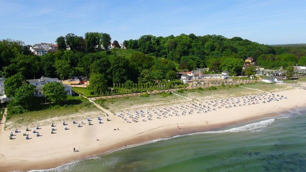A beach with rows of umbrellas and beach chairs, a sandy shore, and a forested hill in the background. - Apartment with sea view, use of swimming pool and sauna - Haus Blick zur See FeWo 01 - (Göhren)