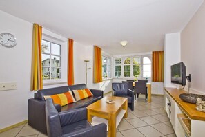 Uma sala de estar moderna com sofá, mesa de jantar, cadeiras, televisão e janelas amplas com cortinas.