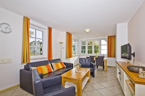 A modern living room with a sofa, dining table, and chairs, a television, and large windows with curtains. - Apartment with a view of the sea, access to the pool and sauna - Haus Blick zur See FeWo 01 - (Göhren)