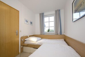A small, tidy room with a single bed, a window with curtains, a bedside lamp, and a wooden door. - Apartment with a view of the sea, access to the pool and sauna - Haus Blick zur See FeWo 15 - (Göhren)