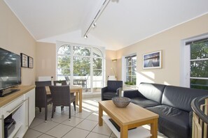 Ein modernes Wohnzimmer mit Flachbildfernseher, Essbereich mit Stühlen und Tisch, einer Couch und großen Fenstern, die für natürliches Licht sorgen.