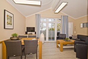 A modern living room with a dining area, a flat-screen TV, and a large window with curtains. - Apartment with balcony, use of swimming pool and sauna - Haus Blick zur See Apartment 14 - (Göhren)