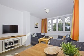 A modern living room with a flat-screen TV, a wooden dining table, a sofa, and a window with curtains. - Apartment with a view of the sea, access to the pool and sauna - Haus Blick zur See FeWo 04 - (Göhren)