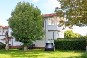 A two-story white house with a red-tiled roof, a large tree in the front yard, and a small table with chairs outside. - Apartment with a view of the sea, access to the pool and sauna - Haus Blick zur See FeWo 04 - (Göhren)