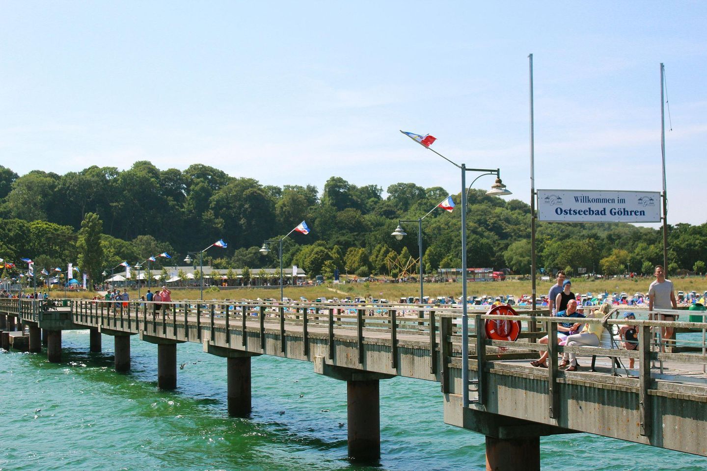 Eine Mole mit einem Schild, das Besucher in Ostseebad Göhren willkommen heißt, mit Blick auf das Wasser und eine bewaldete Gegend im Hintergrund.