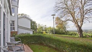 A garden with a paved patio, a white building, and a large tree. - Apartment with terrace, use of swimming pool and sauna - Haus Blick zur See FeWo 13 - (Göhren)