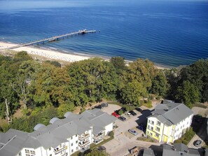 Eine Vogelperspektive zeigt eine Küstenlandschaft mit einem Steg, der sich ins Meer erstreckt, umgeben von dichtem Grün und Wohnhäusern mit Parkplätzen.