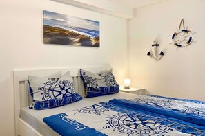 Un dormitorio con temática náutica, que presenta una cama con colcha y almohadillas con motivos de vela en azul y blanco, y un atril de pared con una brújula.