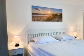 A bedroom with a bed, two bedside lamps, a nightstand, and a wall-mounted picture of a beach scene. - Beachfront Vacation Apartment with Balcony - Strandresidenz Brandenburg FeWo 29 - House (Göhren)