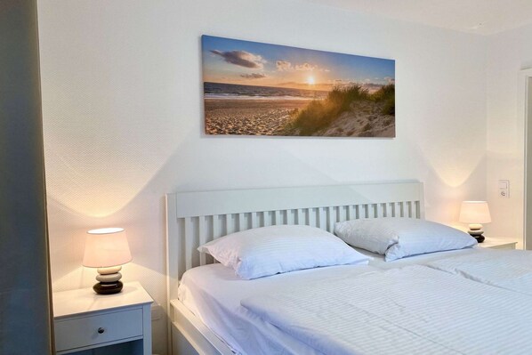 Quarto com cama, duas luminárias de cabeceira, mesa de cabeceira e quadro na parede retratando uma praia.