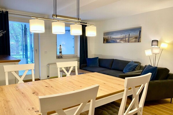 Una sala de estar moderna con una mesa de comedor de madera, sillas blancas, un sofá gris oscuro y un cuadro con una escena de playa.