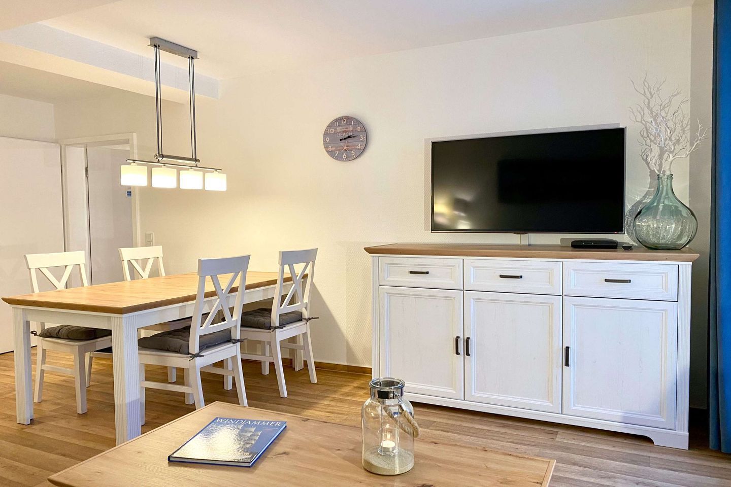 Una sala de estar moderna con una mesa de comedor de madera, sillas blancas, un televisor en un mueble bajo y un reloj en la pared.
